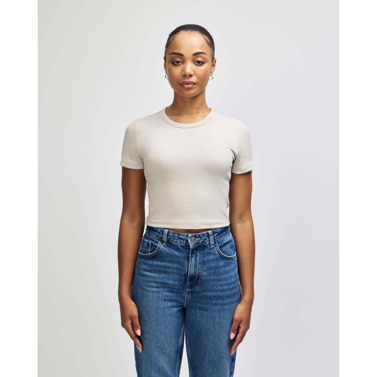 A beige women's crop fitted T-shirt displayed on a model, paired with blue jeans. Features a logo.
