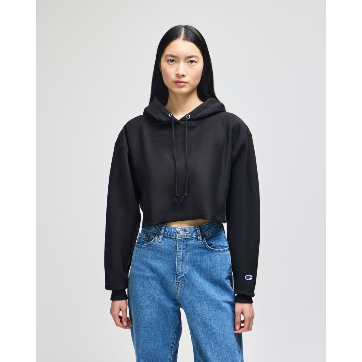 A black cropped hoodie with a hood and long sleeves, worn with blue jeans. It features a logo on one sleeve.