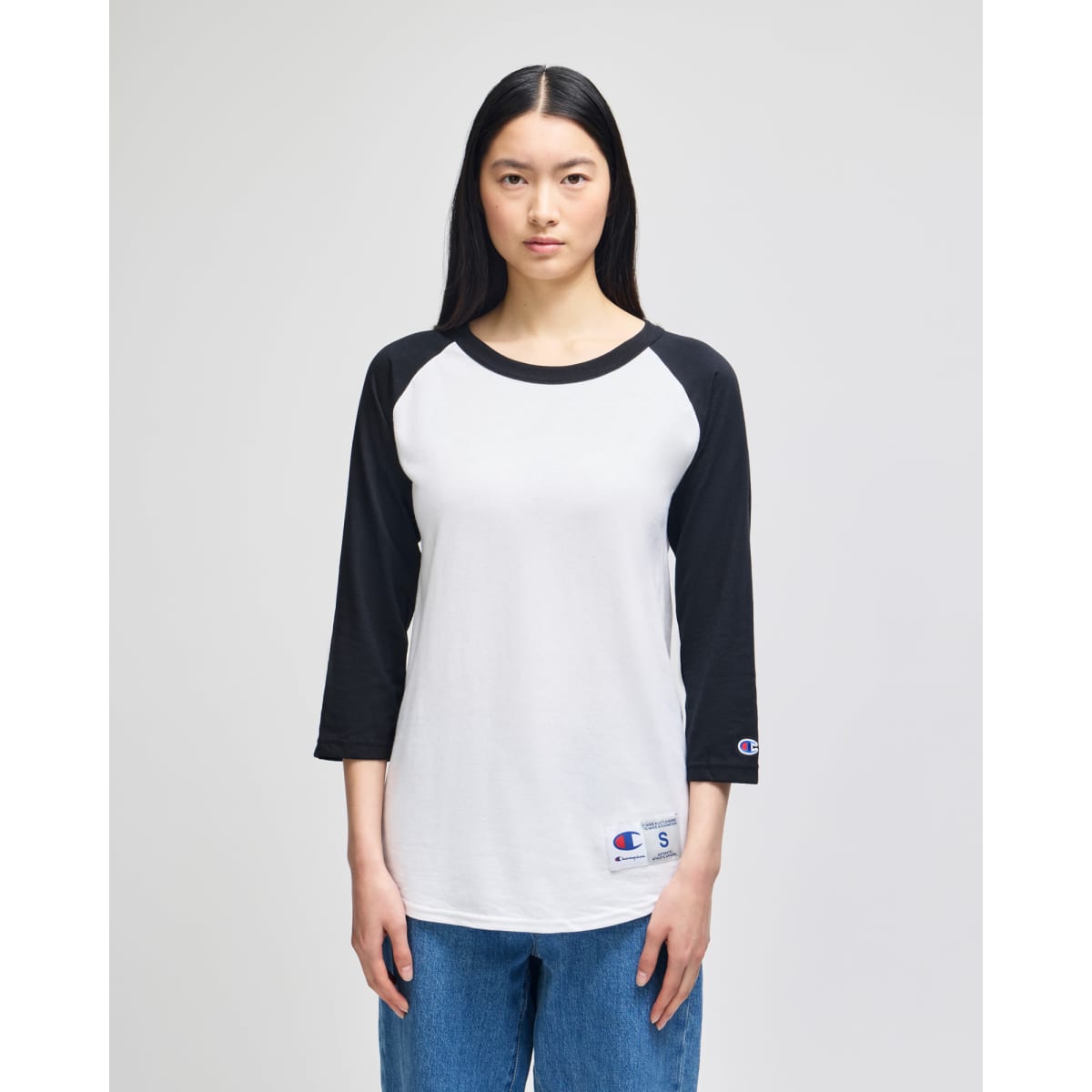 A white and black Champion Adult Raglan Baseball T-Shirt displayed on a model, featuring a logo on the sleeve.