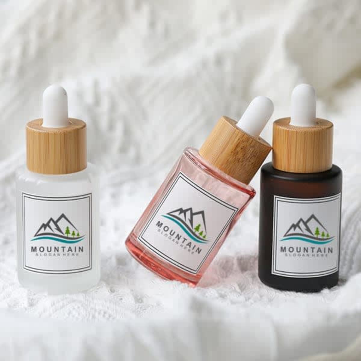 A collection of three 30mL frosted glass dropper bottles in clear, pink, and dark amber with bamboo lids and logos.