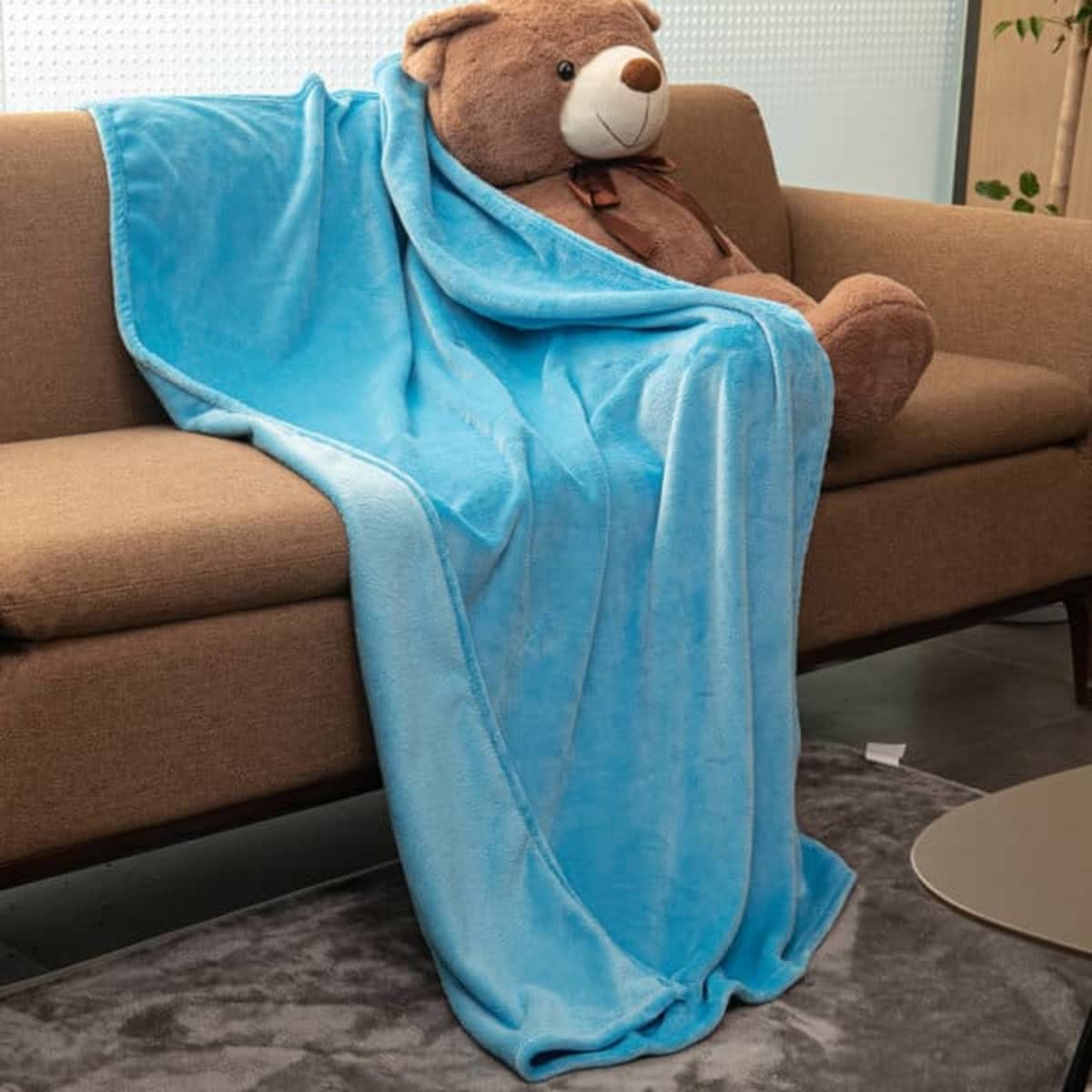 A blue flannel fleece throw blanket draped over a couch, partially covering a large teddy bear.