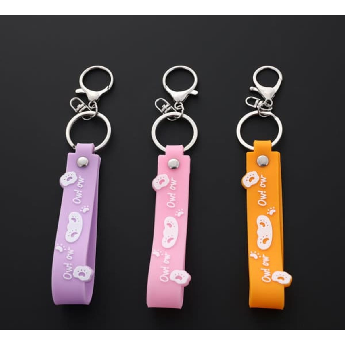 Three colourful PVC soft rubber keychains with lanyards in purple, pink, and orange, featuring small decorations and a logo.