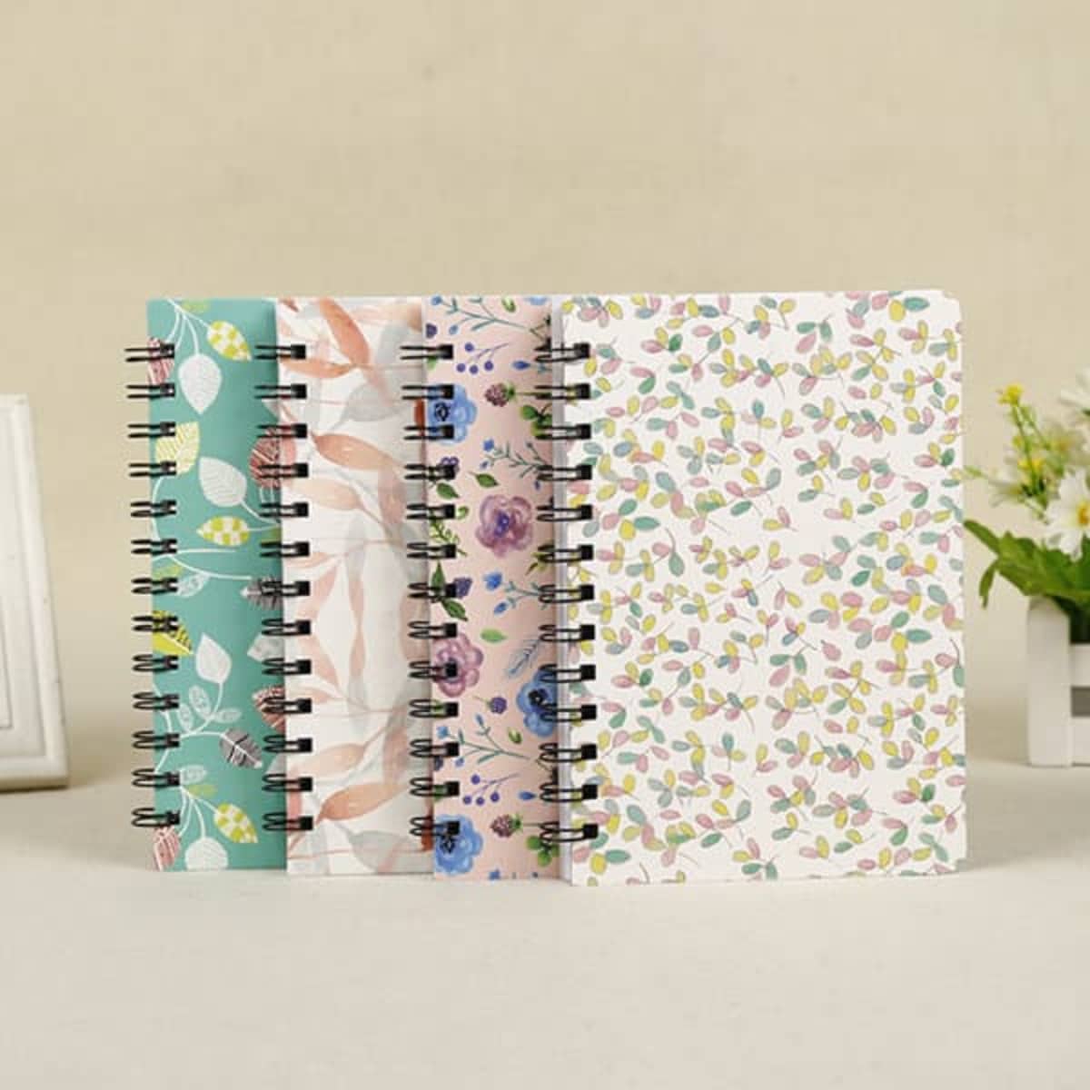 Four spiral-bound hardcover planners in various floral and leaf designs, featuring pastel colours and a logo on the front.