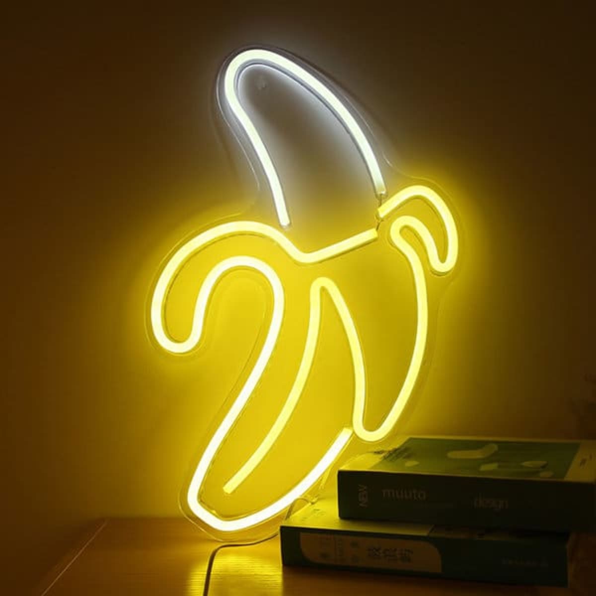A yellow LED neon light shaped like a banana, illuminated against a wall, with books underneath.