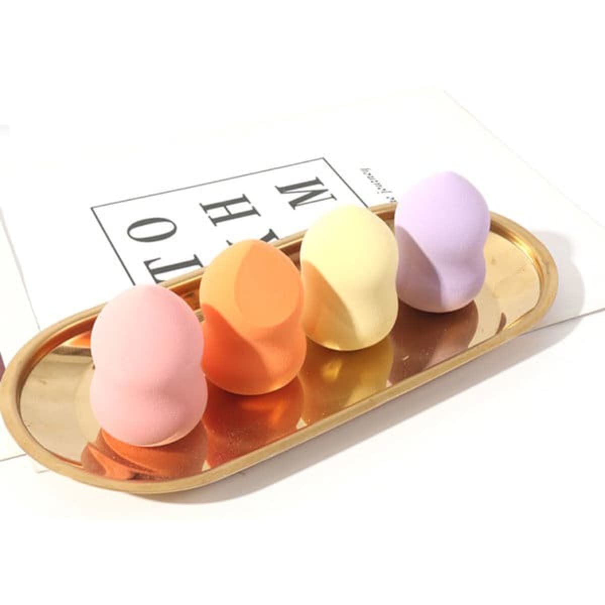 A tray holds four gourd-shaped makeup sponges in pink, orange, yellow, and purple. The tray is gold in colour.