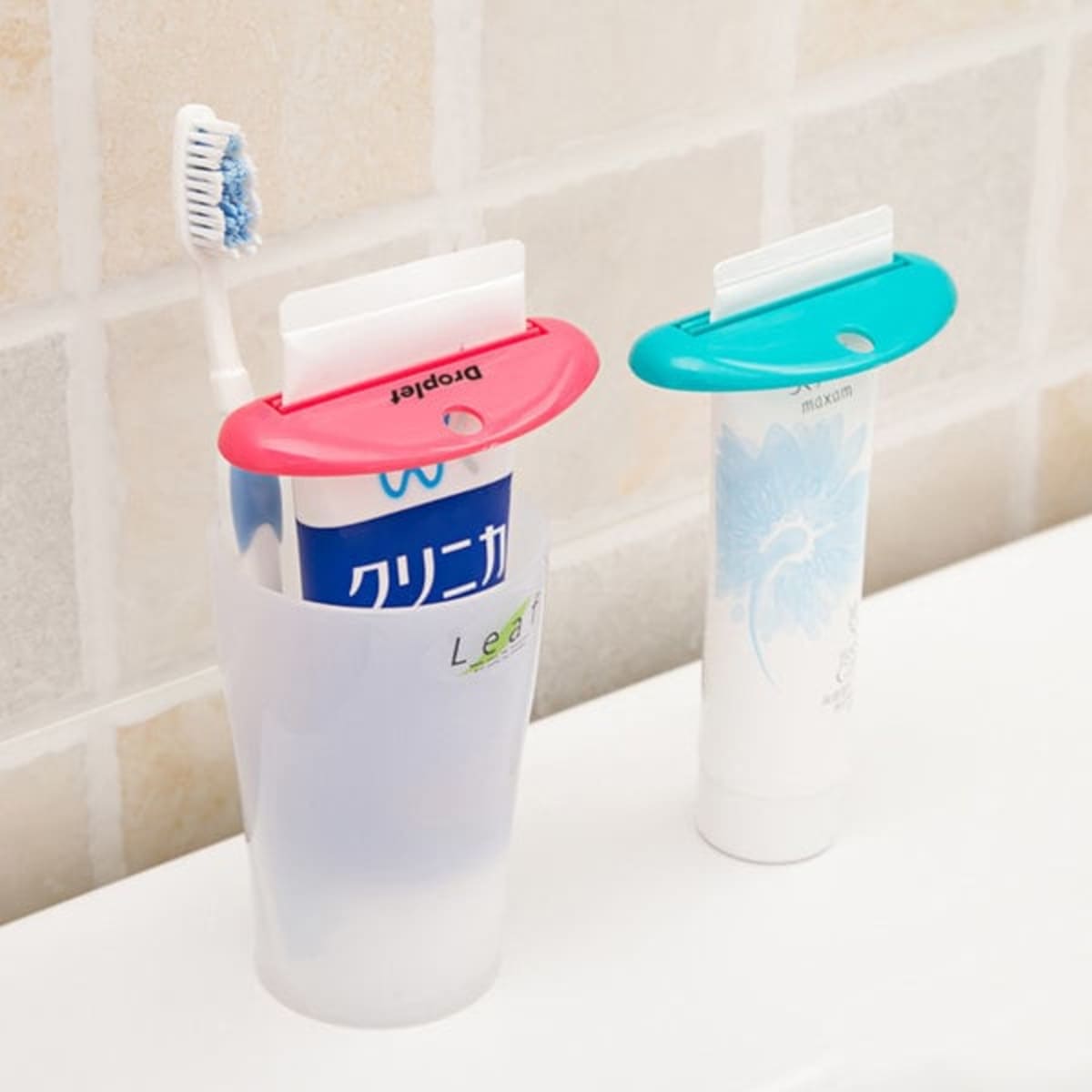 A toothpaste tube squeezer dispenser in pink and blue, alongside a toothbrush, in a bathroom setting.
