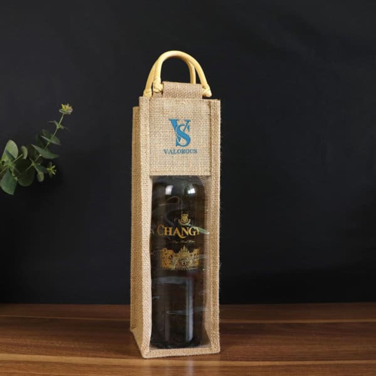 A drink bottle packaged in a natural jute wine bag with a clear window and yellow handle. Features a logo on the front.
