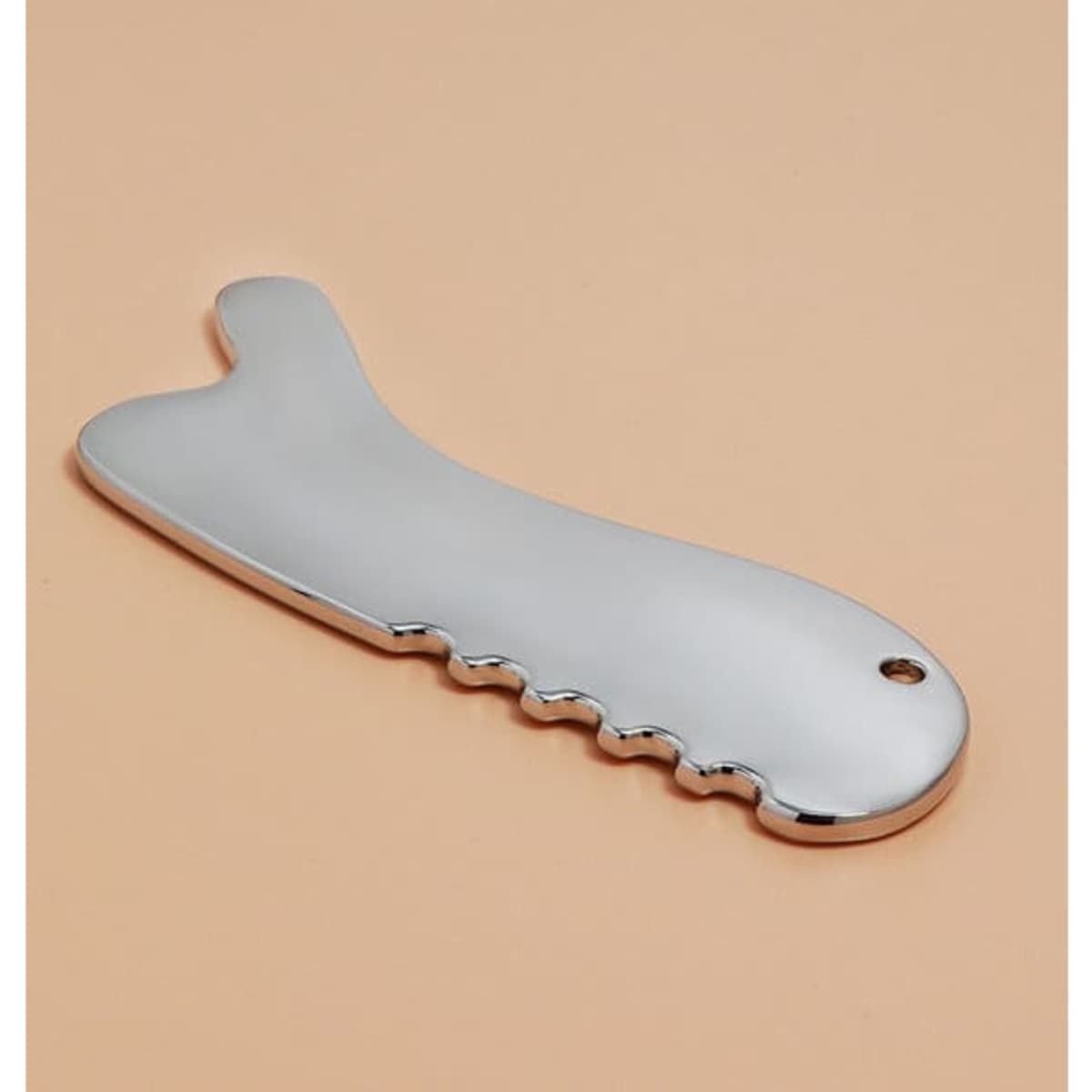 A silver multifunctional metal Gua Sha massage tool with a textured grip and rounded edges, set against a beige background.