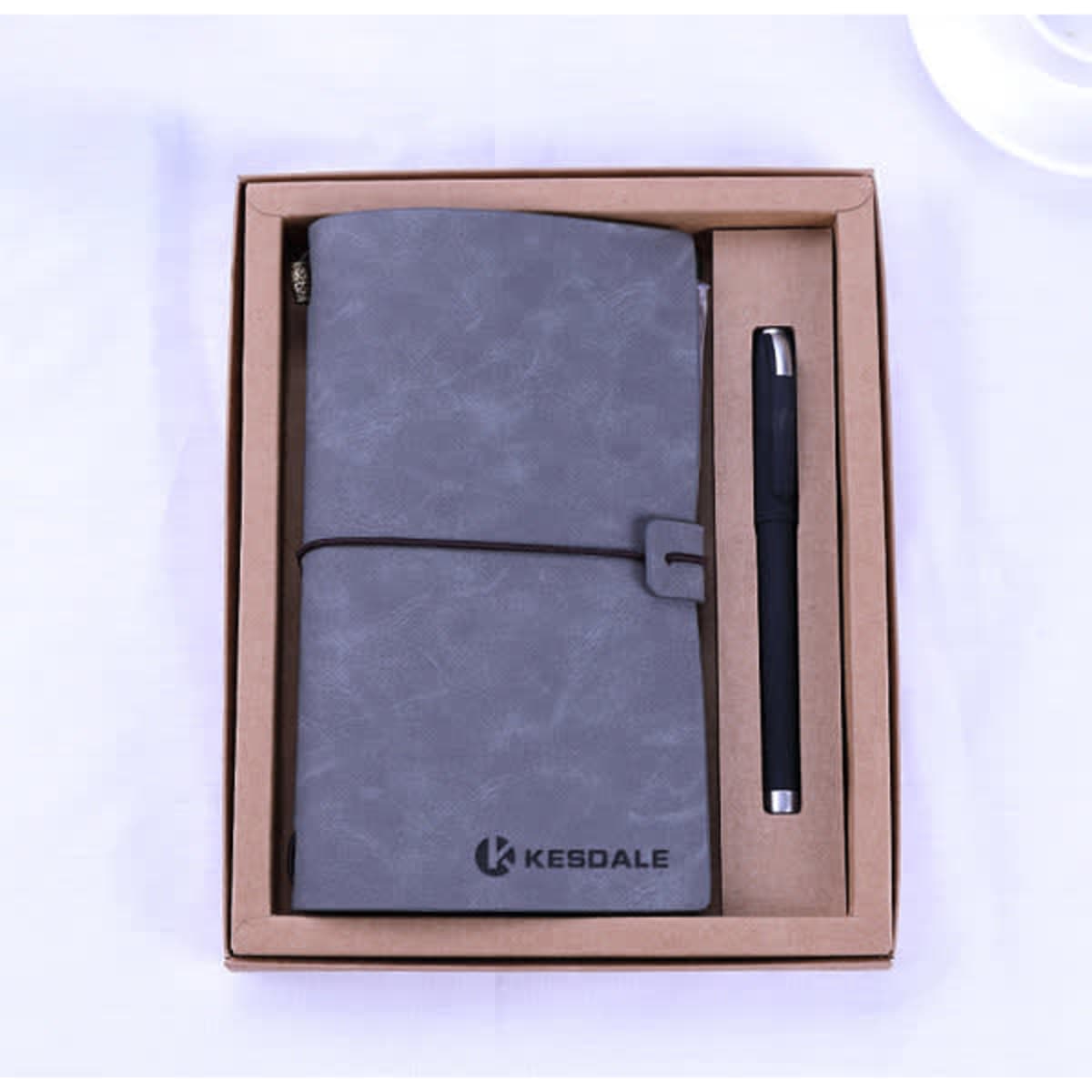 A grey retro travel notebook and pen displayed in a brown gift box, featuring a logo.