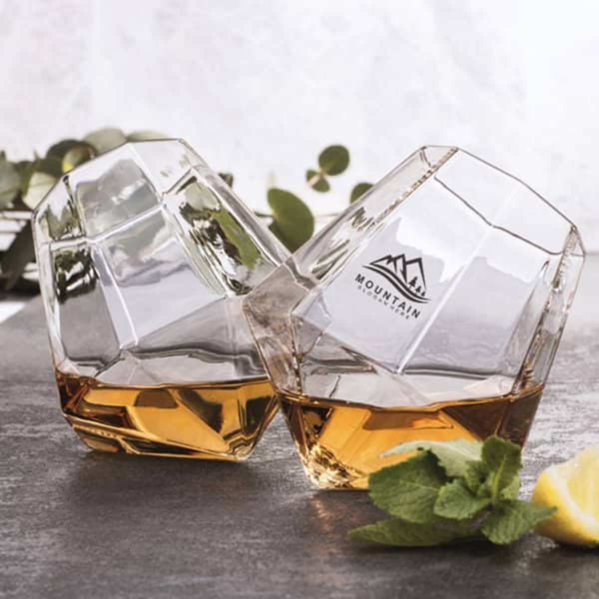 Two diamond-shaped stemless wine glasses in clear glass, featuring a logo, filled with a brown liquid.