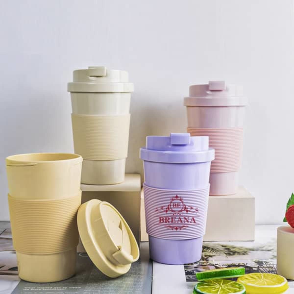 A set of four biodegradable plastic travel coffee cups in cream, pastel yellow, light purple, and pink, featuring lids.