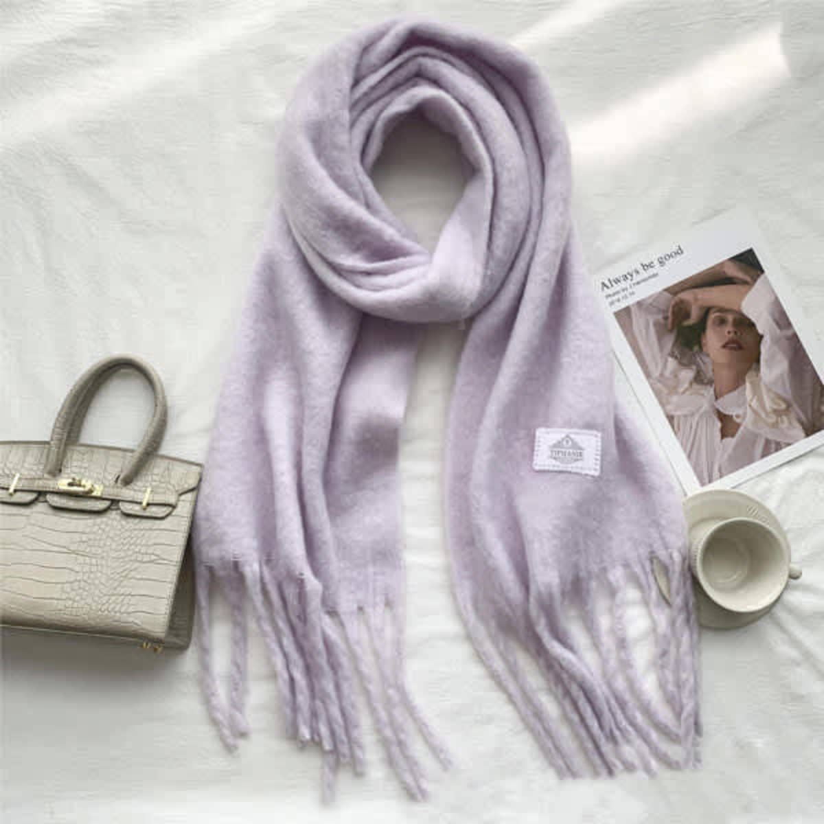 A lilac wool scarf with tassels, featuring a small logo, laid out beside a handbag and a magazine.