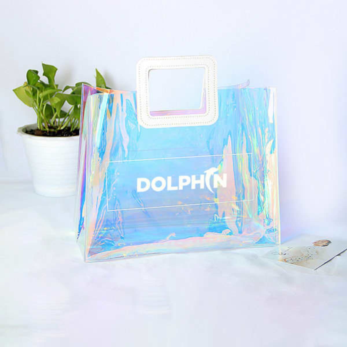 A clear, iridescent tote bag with a white PU handle and a branding logo, beside a small potted plant.