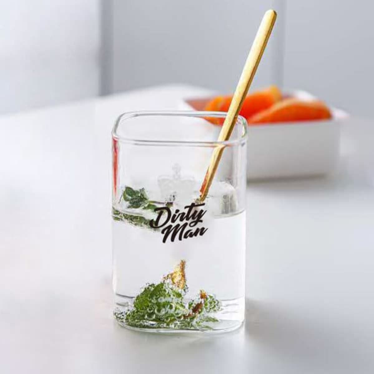 A square 9 oz. clear glass water cup filled with water and mint leaves, featuring a logo and a gold stirring stick.