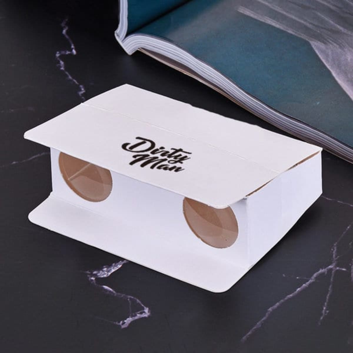 A custom folding paper binocular in white with circular lenses and a logo. The background features a magazine.