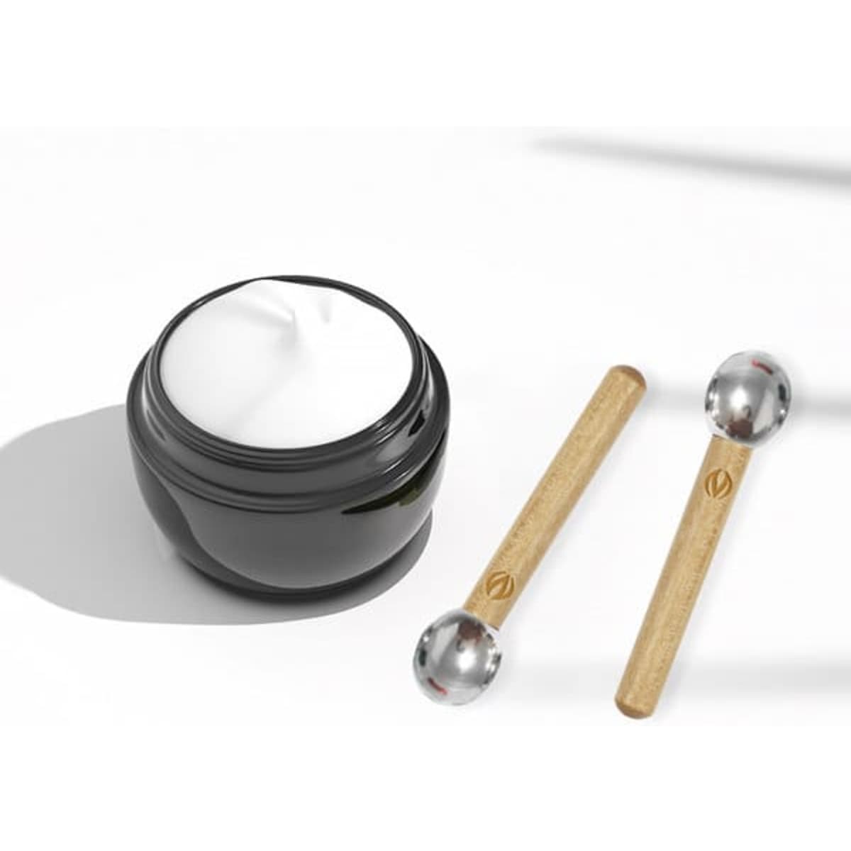A black jar of cream with two wood handle metal eye roller massage sticks beside it.