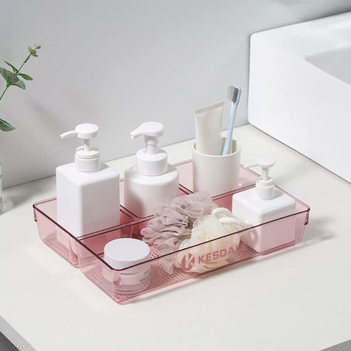A pink compartment cosmetic tray featuring various white containers and accessories, set on a bathroom countertop.
