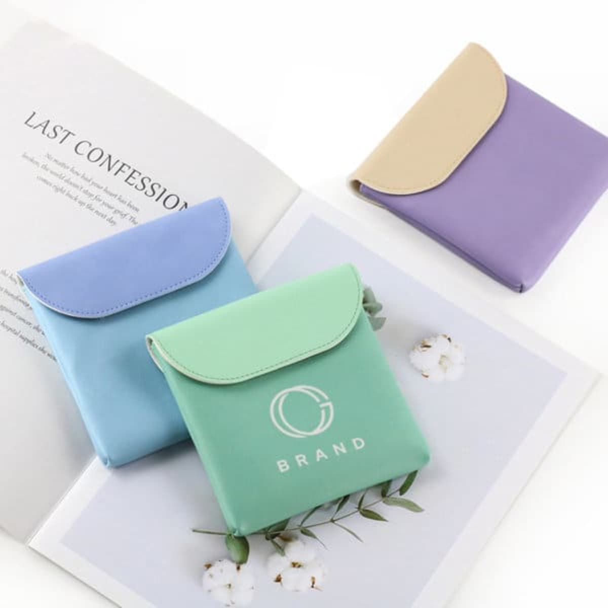 Three portable coin purses in light blue, mint green, and lilac, arranged on a floral background. One purse features a logo.