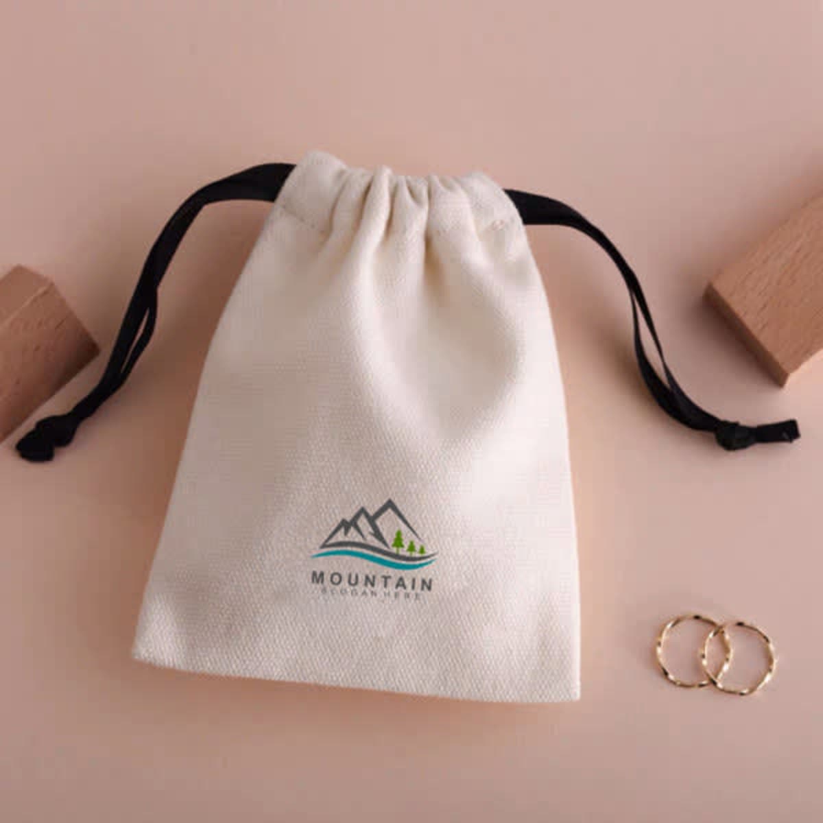 A cream drawstring canvas bag with black cords and a logo, displayed alongside two gold rings.
