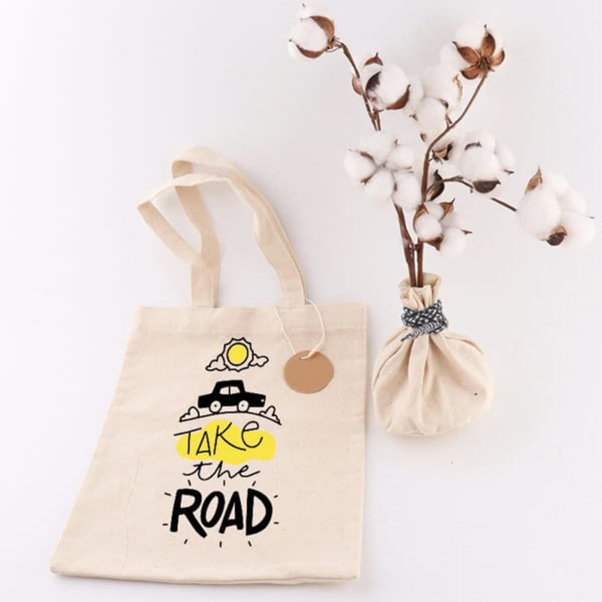A natural canvas tote bag with a design featuring a car, cloud, and sun, alongside a vase of cotton stems.
