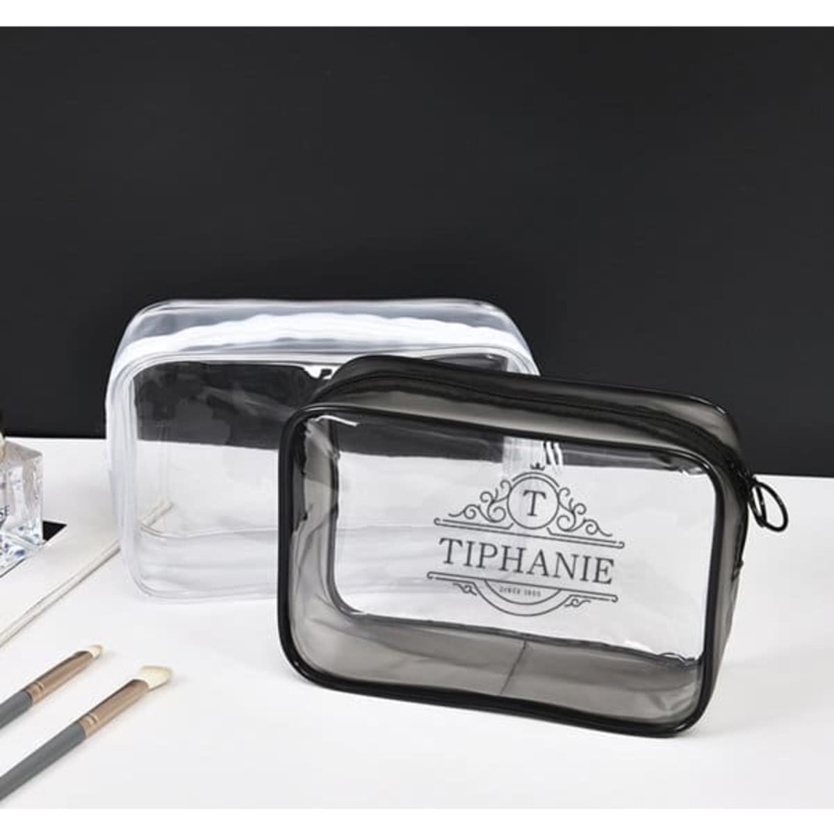 Two large clear PVC zippered toiletry bags in black and transparent, featuring a logo. Makeup brushes are placed beside them.