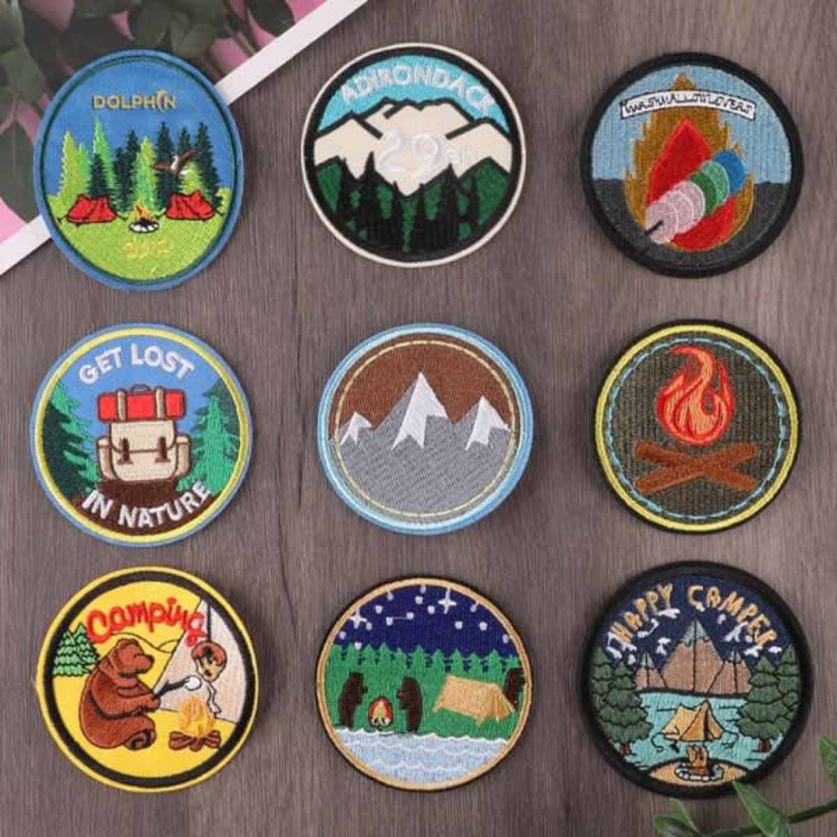 A collection of nine custom embroidered patches featuring various outdoor-themed designs and colours on a wooden surface.