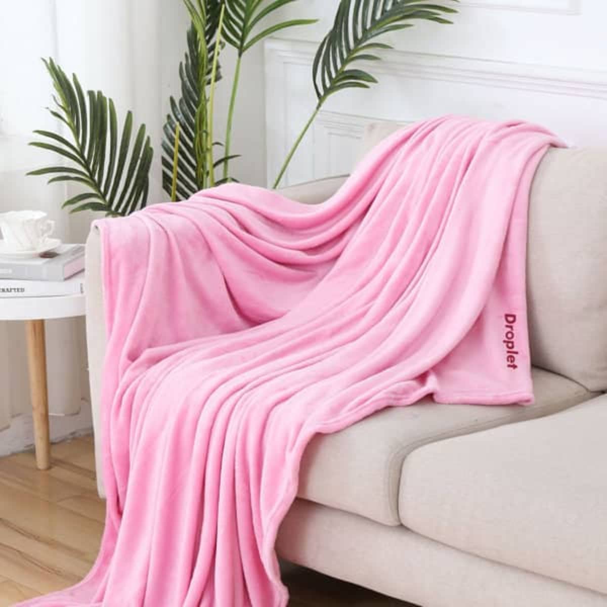 A soft pink flannel fleece blanket draped over a light-coloured sofa, with greenery in the background.