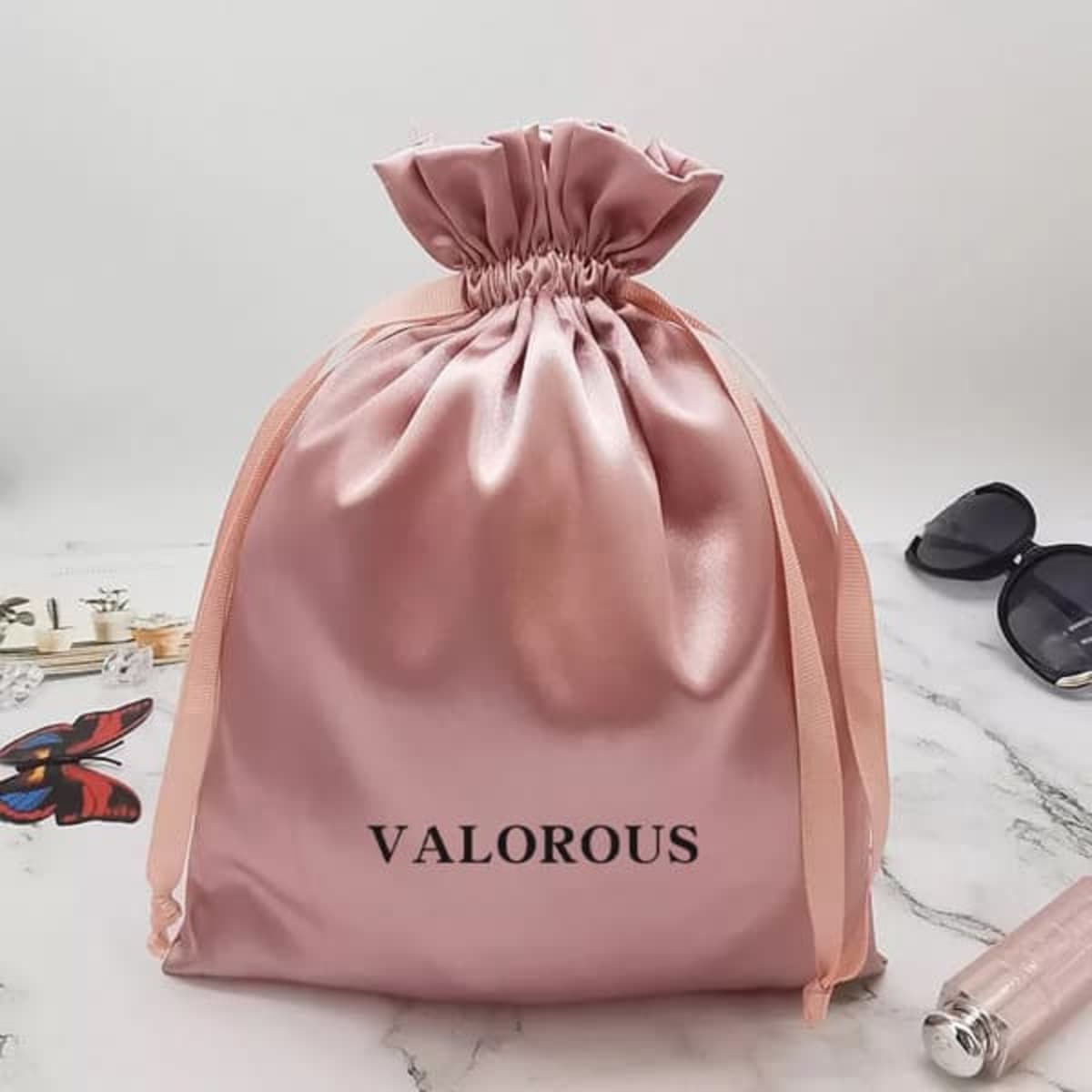 A satin pink drawstring gift bag with a gathered top and ribbon ties, featuring a logo.