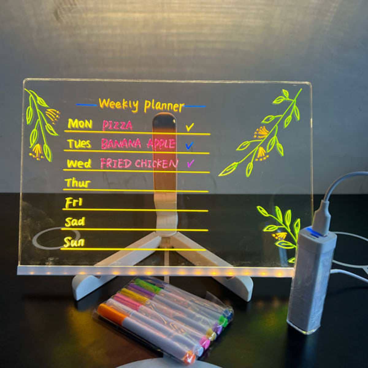 A LED acrylic message board with a colourful weekly planner and markers, featuring floral designs and a charging base.