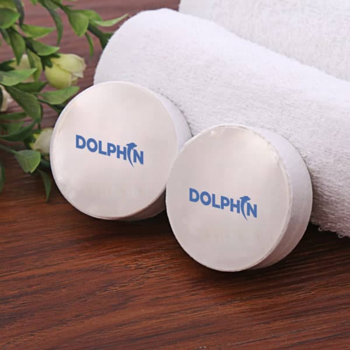 Two round white compressed towels with a logo on them, placed on a wooden surface alongside a rolled white towel and leaves.