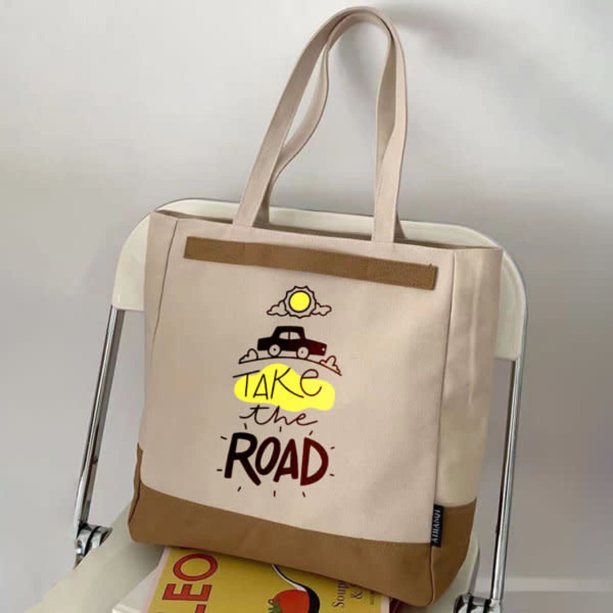 A cream canvas tote bag with a brown base and a graphic design featuring a car and sun, complete with a zipper.
