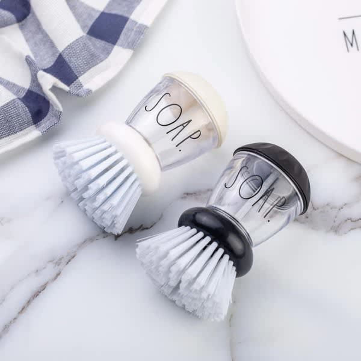 A small soap dispensing palm brush in white and black, featuring bristles and a transparent soap chamber.
