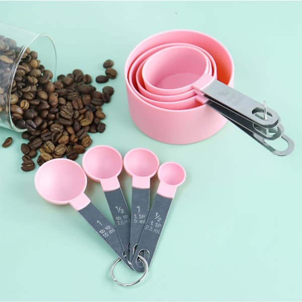A set of eight stainless steel measuring spoons with pink plastic cups, alongside scattered coffee beans.