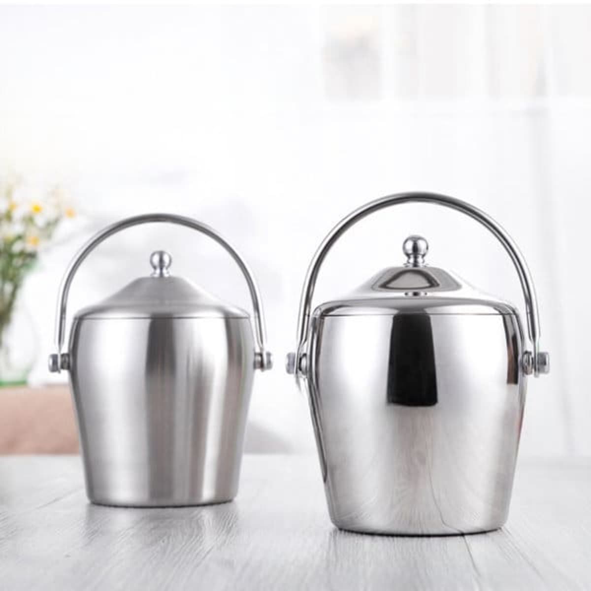 Two stainless steel ice buckets with lids, featuring polished finishes and metal handles, set on a wooden surface.