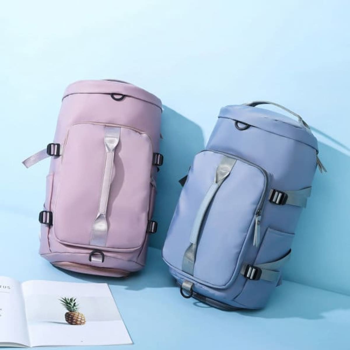 Two gym backpacks in pink and blue, featuring various compartments and straps, alongside an open magazine.
