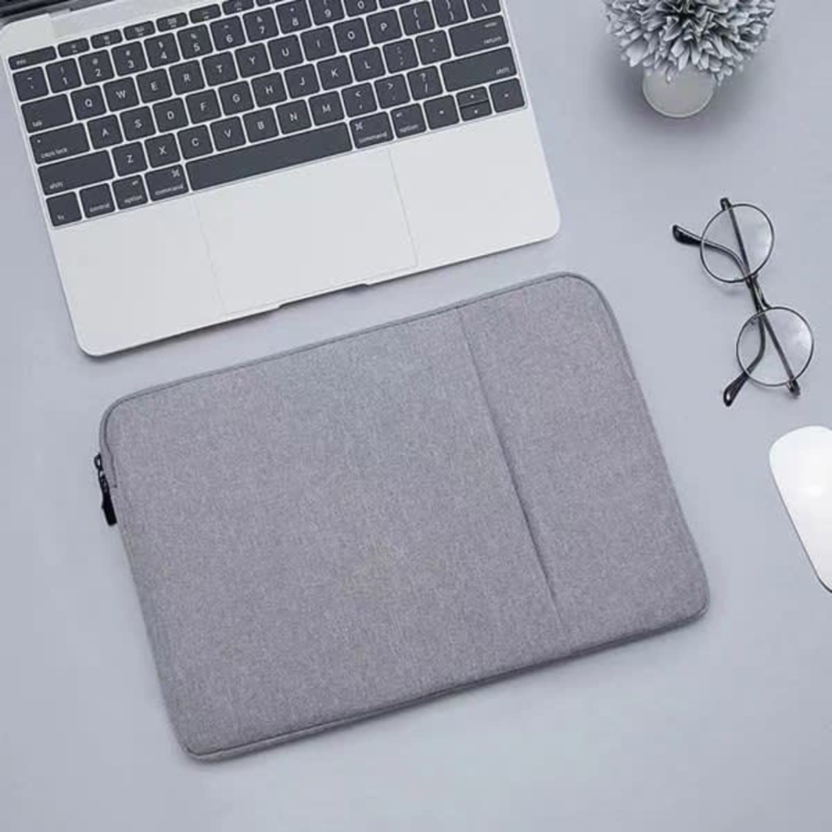 A grey 15.6-inch laptop sleeve case is displayed on a desk beside a laptop, glasses, and a small plant.