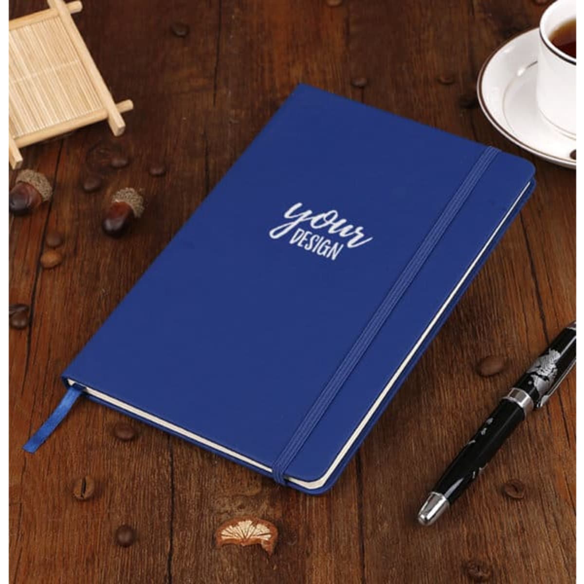 A blue PU hardcover notebook with a strap, placed on a wooden surface beside a coffee cup and a pen.
