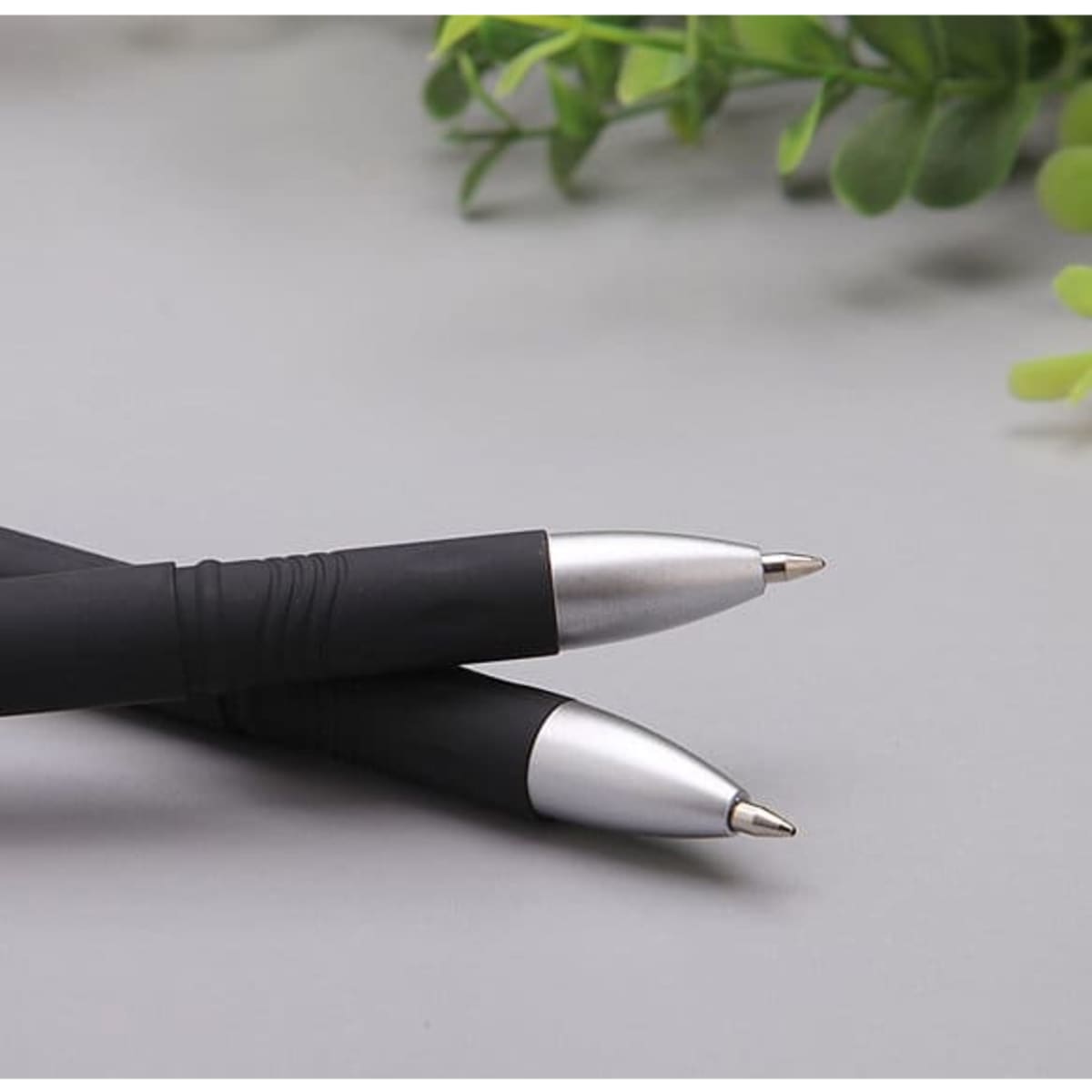 Two black promotional pens with silver accents lying on a grey surface, surrounded by green foliage.