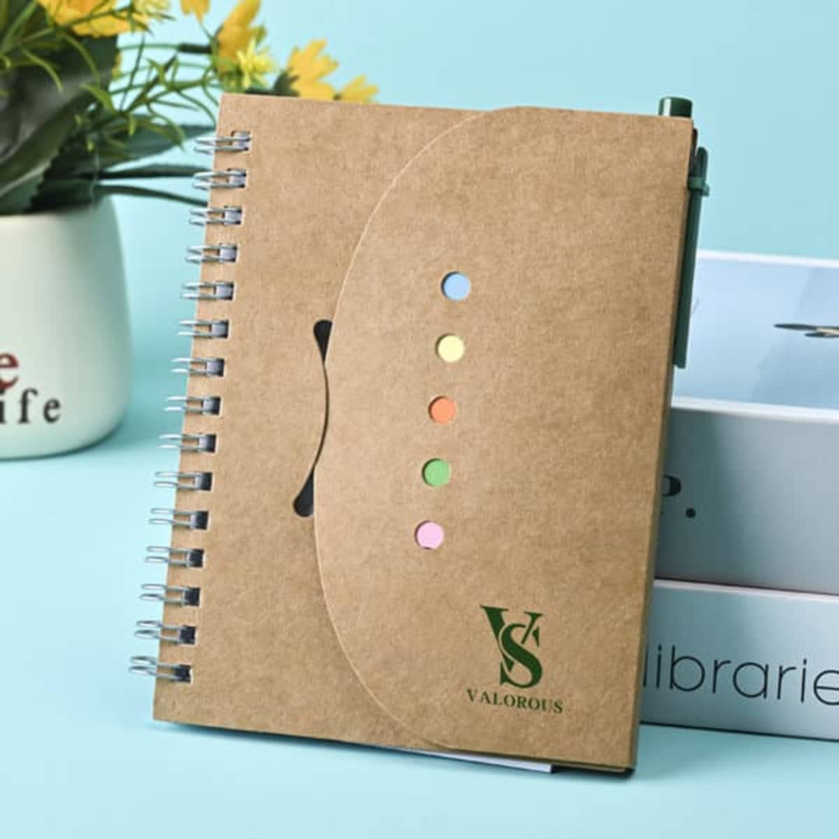 A brown spiral notebook with colourful sticky notes and a pen, featuring a logo on the cover.