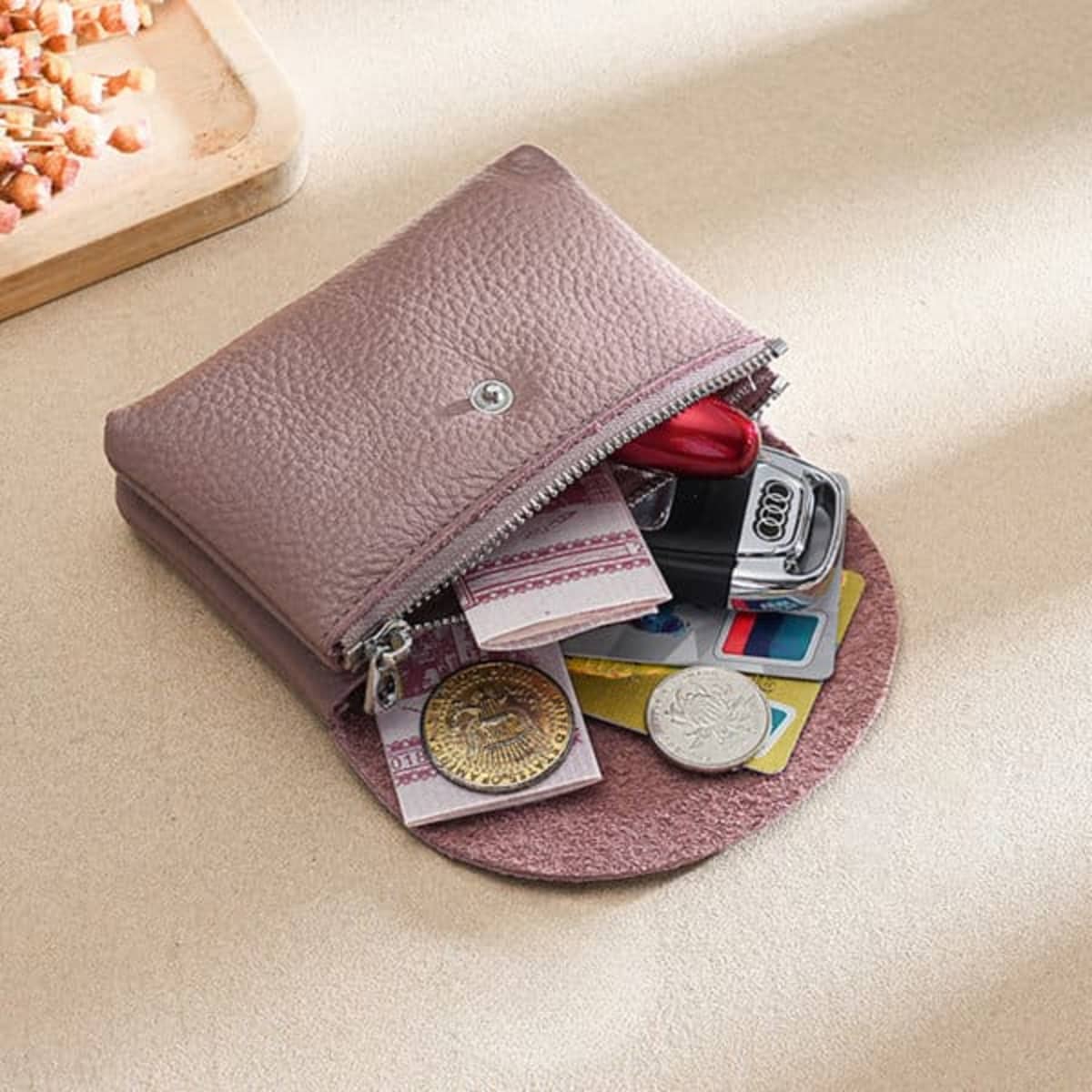 A small leather coin purse in mauve, containing coins, cards, and a key, placed on a textured surface.