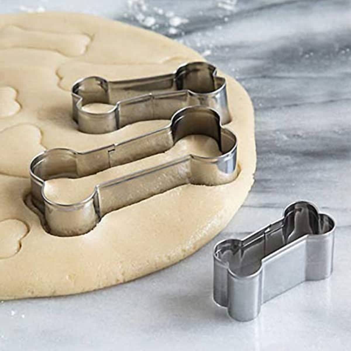 A set of three stainless steel dog bone cookie cutters rests on rolled out dough on a marble surface.