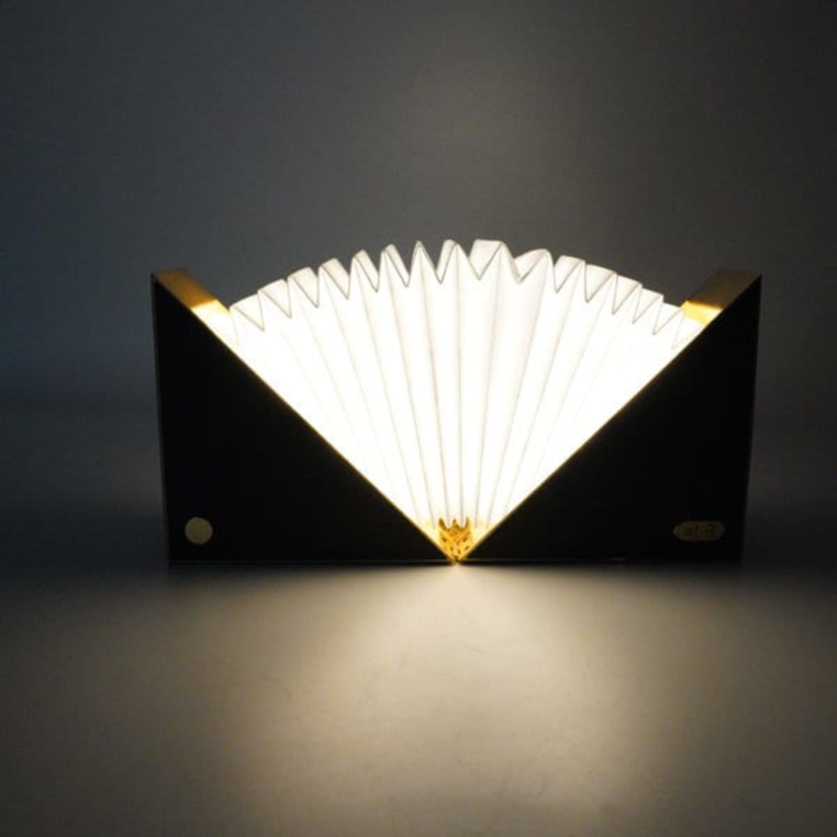 A folding book light with a black exterior and white, pleated interior, featuring gold accents. It has a logo on the side.