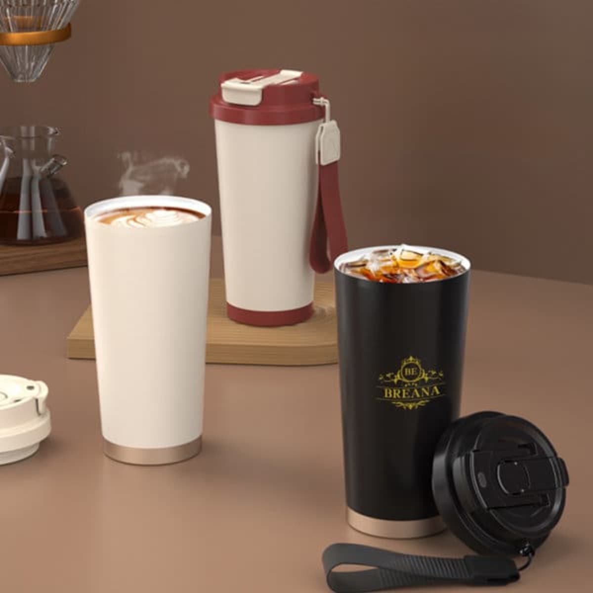 Two stainless steel coffee cups are displayed: one white with a beige base and a red lid, and one black with a gold logo and a black lid.