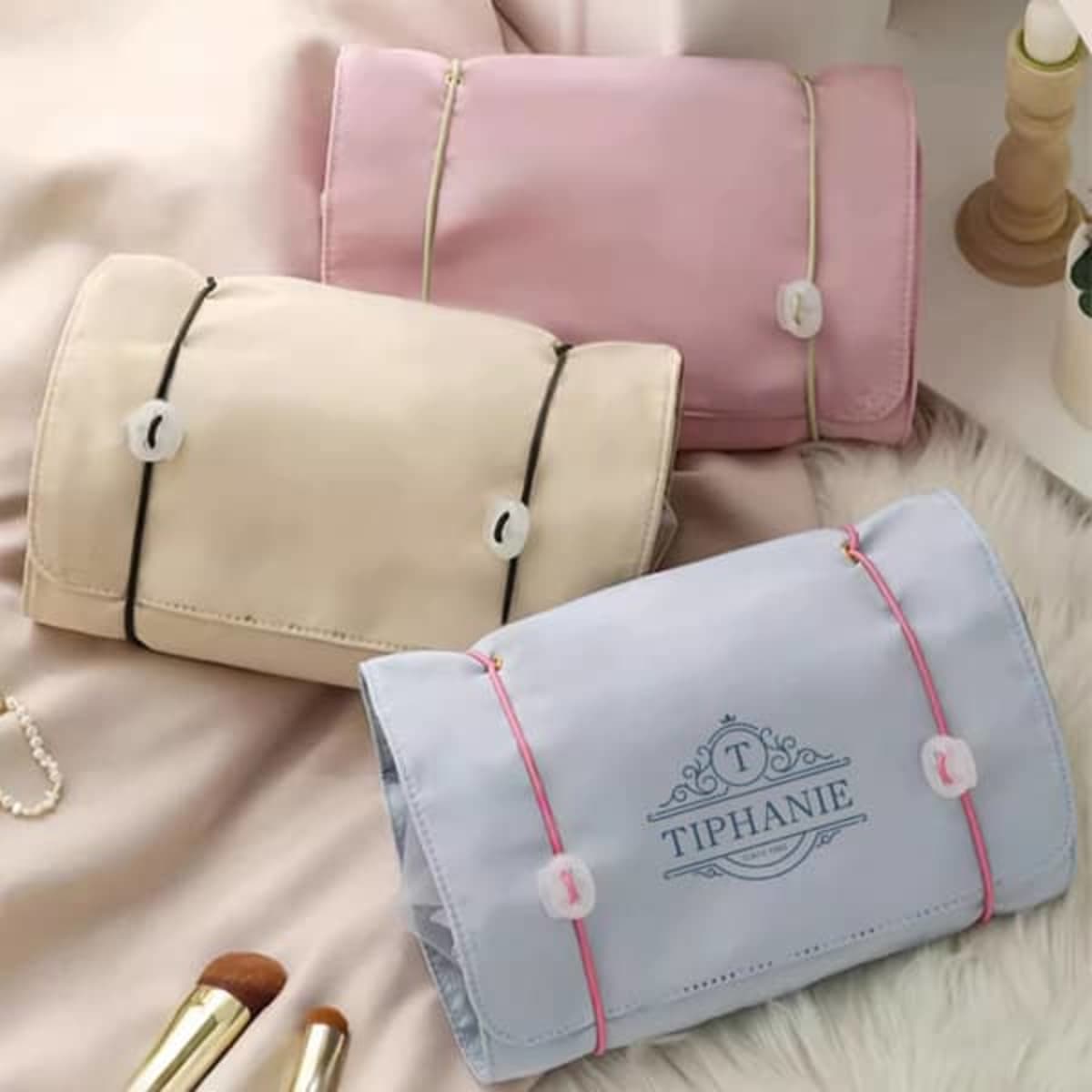 Three folding travel cosmetic bags in pink, cream, and blue, featuring a decorative clasp and a logo.
