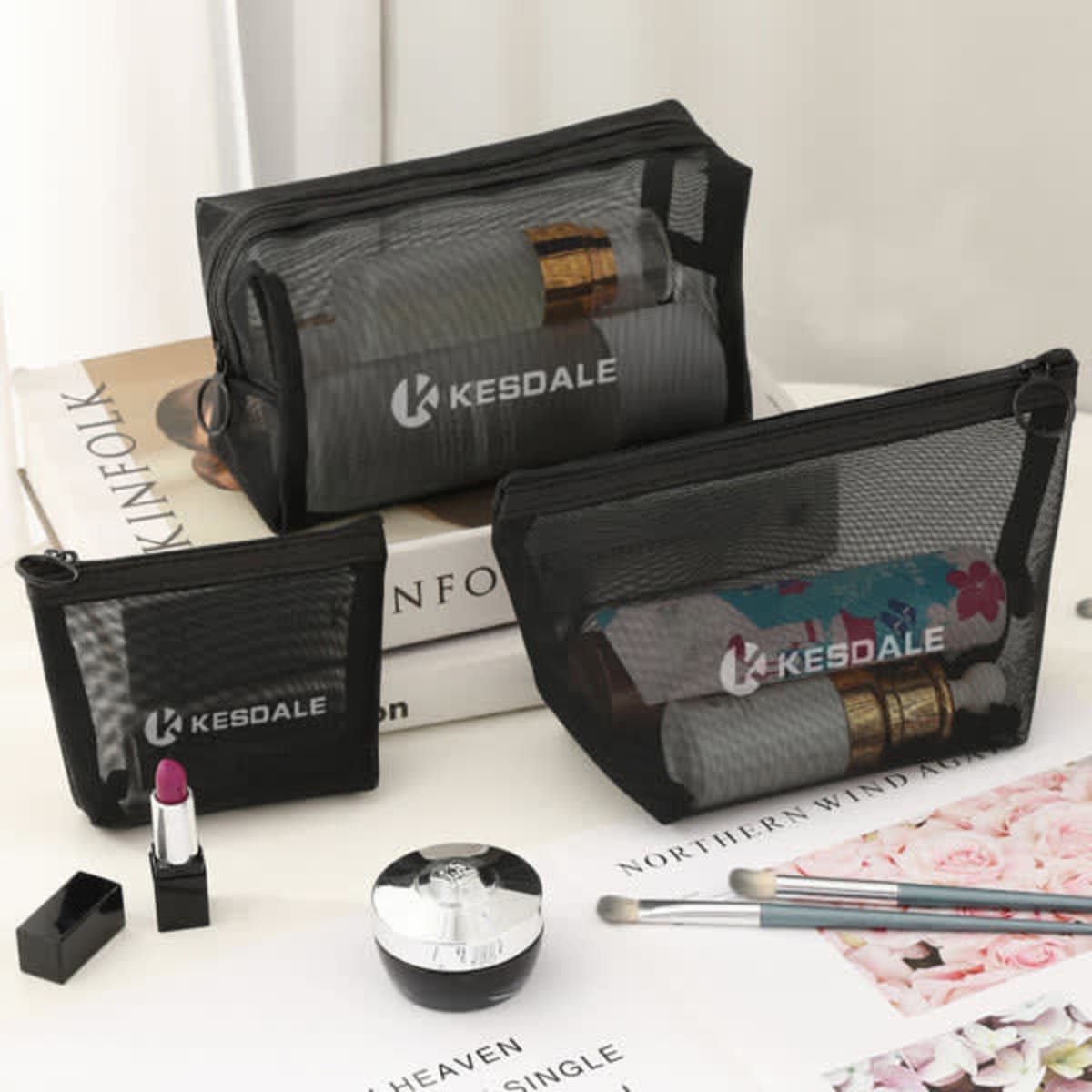 Three black mesh cosmetic bags of varying sizes displayed on a table with beauty products and a magazine.
