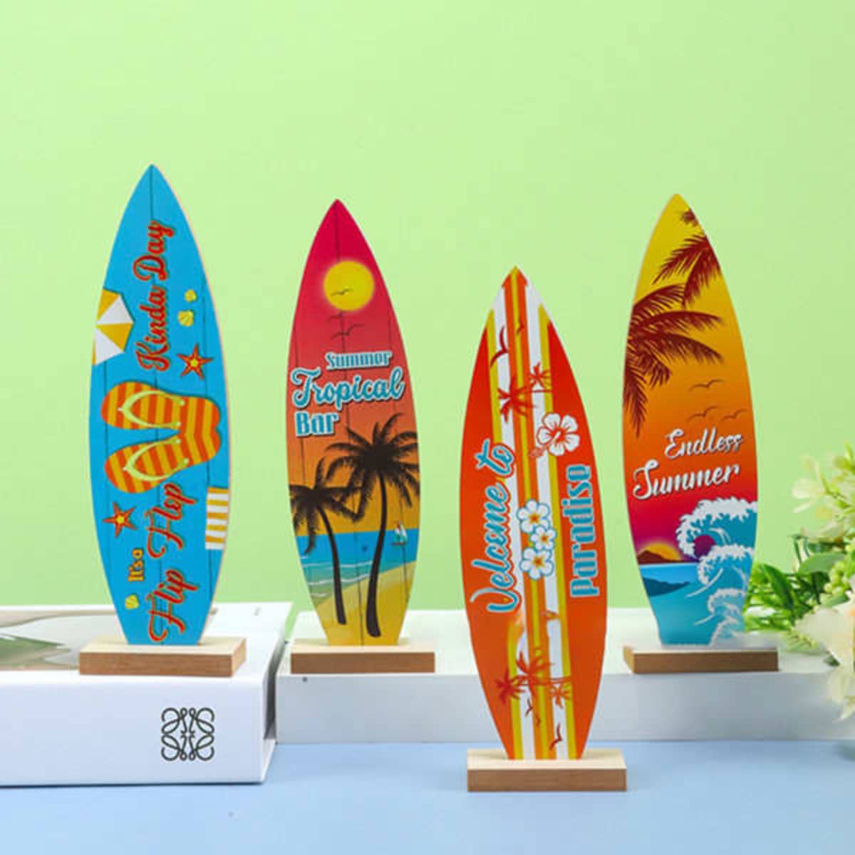 A collection of four colourful surfboard ornaments featuring tropical designs, displayed on wooden bases against a green background.