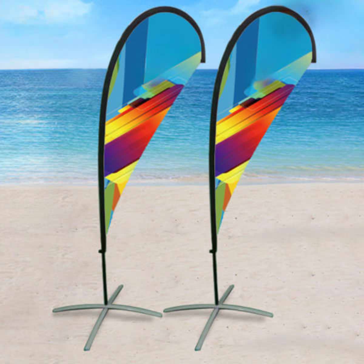 Two vibrant, multicoloured advertising flag banners on stands beside a beach with ocean background.