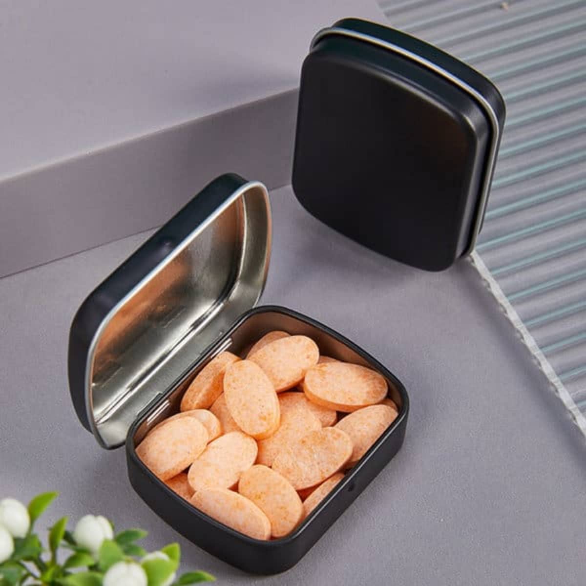 A black mini metal hinged box container open to reveal orange oval-shaped candies inside.