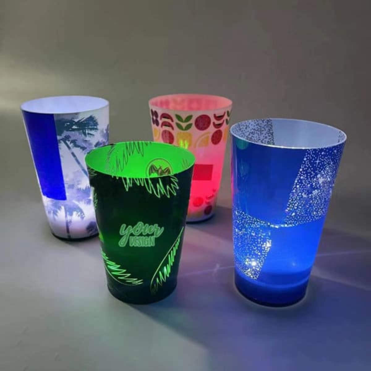 Four illuminated party cups in various designs and colours: green, blue, pink, and a patterned cup with a logo.