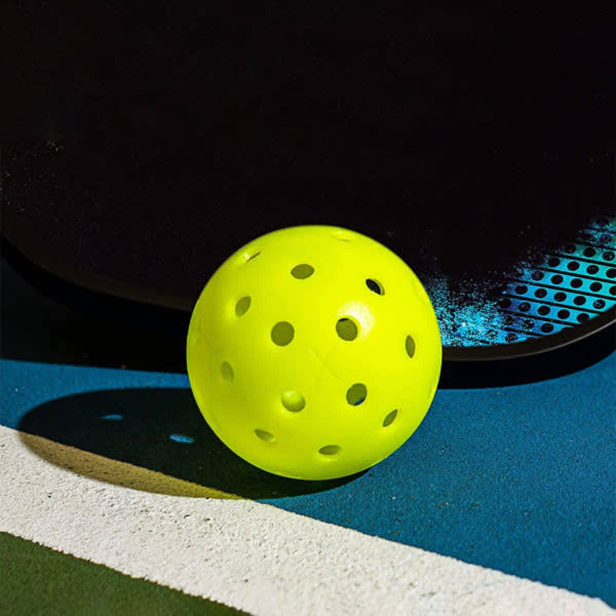 A bright yellow pickleball with holes sits on a blue and white surface next to a paddle with a textured design.